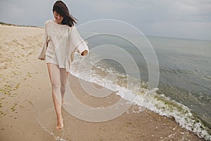 Carefree beautiful woman with windy hair running on sandy beach at cold sea waves, having fun. Stylish young happy female in