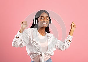 Carefree black woman in headphones dacing to her favorite music with closed eyes on pink studio background