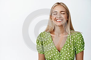Carefree blond girl close her eyes, smiles and feels happy, breathing, standing in green dress over white background