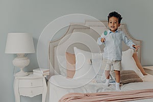 Carefree cute afro american child boy with lollipop jumping ob bed at home