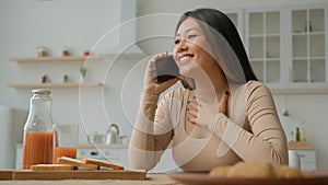 Carefree ethnic Asian woman housewife talking mobile phone in kitchen talk with friend calling speak cellphone share