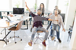 Carefree excited diverse workers having fun riding oh chairs celebrating friday together. Happy employees enjoy funny competition