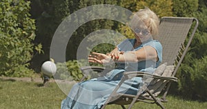 Carefree female retiree sitting in garden chair and applying hand moisturizer. Portrait of happy relaxed Caucasian lady