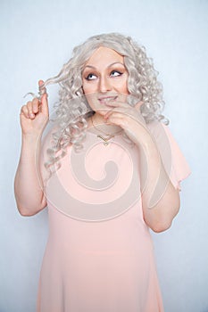 Carefree girl in a pink dress twists her blonde curly hair on her finger and rejoices on a white monochrome Studio background