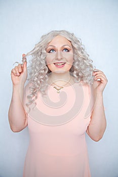 Carefree girl in a pink dress twists her blonde curly hair on her finger and rejoices on a white monochrome Studio background