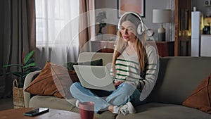 Carefree girl surfing laptop listening music in wireless headphones at couch.