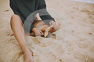 Carefree hipster girl holding herb and sitting on beach, cropped view on tanned legs. Stylish boho woman in modern swimsuit and