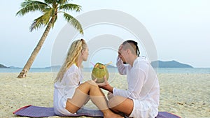 Carefree loving couple facing each other sits in tropical beach under palm tree and drink coconat cocktail, kissing and