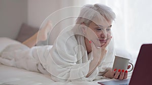 Carefree mature woman smiling lying on bed watching online movie on laptop. Portrait of happy relaxed positive Caucasian