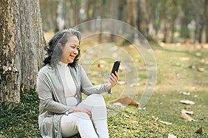 Carefree middle aged woman using smart phone under tree in the park at sunny beautiful day. Elderly lifestyle and