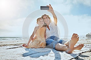 Carefree mother and daughter taking a selfie while sitting on the beach. Happy little girl and grandmother smiling while