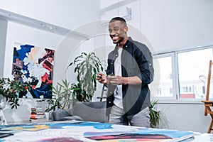 Carefree perky black man standing next to a big canvas with painting on a table