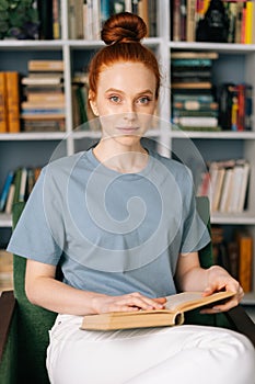 Carefree redhead young woman student is reading book enjoys of rest at home office, looking at camera.