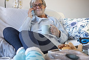 Carefree senior woman relax in sofa at home using smartphone and having a break with food and drink
