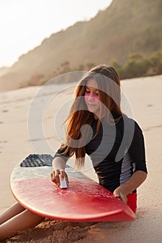 Carefree sporty woman waxes surface of surfboard with piece of wax, dressed in wetsuit, sits at sandy beach alone