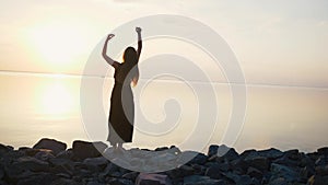 Carefree woman in long black dress dancing on beach at sunset