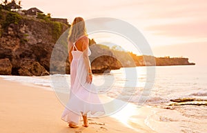 Carefree woman in white dress walking on the beach at sunset