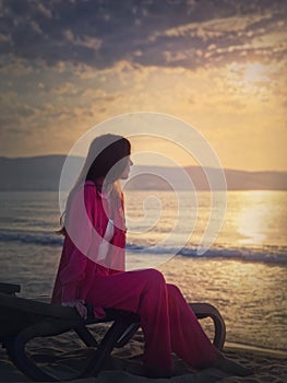 Carefree young woman aesthetic portrait sitting relaxed on the sunbed at the beach looking at the sunrise above the sea. Beautiful