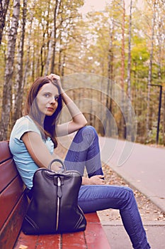 Carefree young woman in blue pants sitting on bench in park. Girl in a pensive mood posing on an autumn day. Fall concept