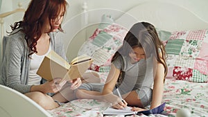 Careful mother helping her little cute daughter with homework for elementary school. Loving mom reading a book and girl
