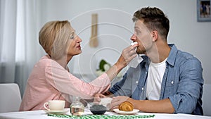 Careful mother wiping adult sons mouth during having tea at home, overprotection