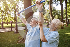 Careful woman helps senior man to do exercise with bar in park