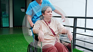 Caregiver asian woman walking while care elderly patient sitting on wheelchair in garden at hospital.