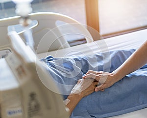 Caregiver and elderly senior patient (aged old adult person) holding hands in hospital bed or nursing hospice
