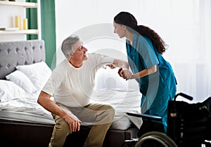 Caregiver helping elderly male to get into wheelchair at home. Rehabilitation and medical care for disabled seniors