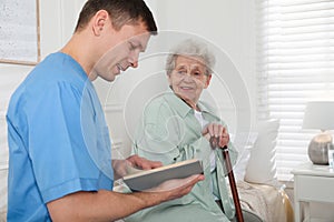 Caregiver reading book to senior woman indoors. Home health care service