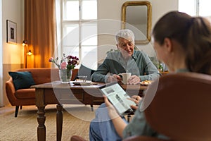 Caregiver reading online newspaper in digital tablet during taking care of senior man.