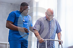 Caregiver is teaching old man to walk with walker. Professional nurse and patient in a nursing home. Assistance