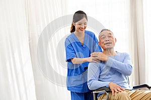 The caregiver therapist stands with an Asian senior sitting in a wheelchair and touches their hands on senior shoulder together.