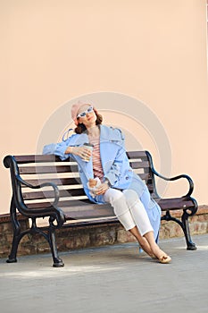 Careless woman in beret and blouse, raincoat and sunglasses sitting on bench with snack