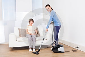 Caretaker cleaning floor while senior woman sitting on sofa