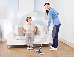 Caretaker cleaning floor while woman sitting on sofa