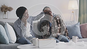 Caring african american woman calling to doctor, touching forehead of her sick husband, sitting on coach at home