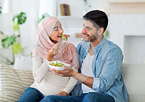 Caring arab man giving his young pregnant wife fresh vegetable salad, sitting on sofa in living room at home