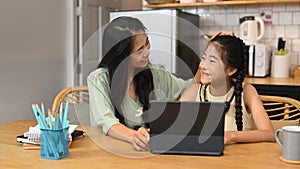 Caring asian mom helping her daughter doing homework, studying online in kitchen