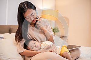 A caring Asian mom is putting her little son to sleep in her arms while she is talking on the phone