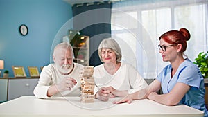 caring for elderly, joyful female and male pensioners have fun playing board game with a nurse sitting at table in room