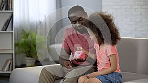 Caring father teaching child to save money, girl putting coins into piggy bank