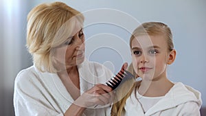 Caring grandmother combing granddaughter hair in front of mirror, family love