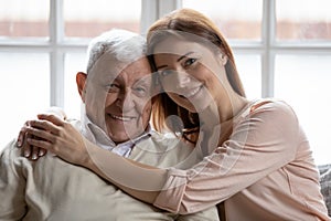 Caring grownup granddaughter hugs elderly 75s grandfather seated on sofa