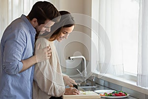 Caring millennial husband hugging shoulders of beloved wife preparing breakfast