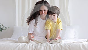 Caring mom sit on bed with preschool kid read book or show image to small boy explaining teaching
