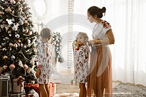 Caring mother braids her little daughter`s braid while second daughter decorates a New Year`s tree in the light cozy