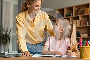 Caring mother checking her little daughter doing homework, cheerful mom and cute female child sitting at table