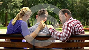 Caring mother and dad supporting sad teen son sitting on bench in park, crisis