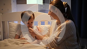 Caring mother measuring temperature to her sick little son lying in bed at night. Concept of children illness, disease